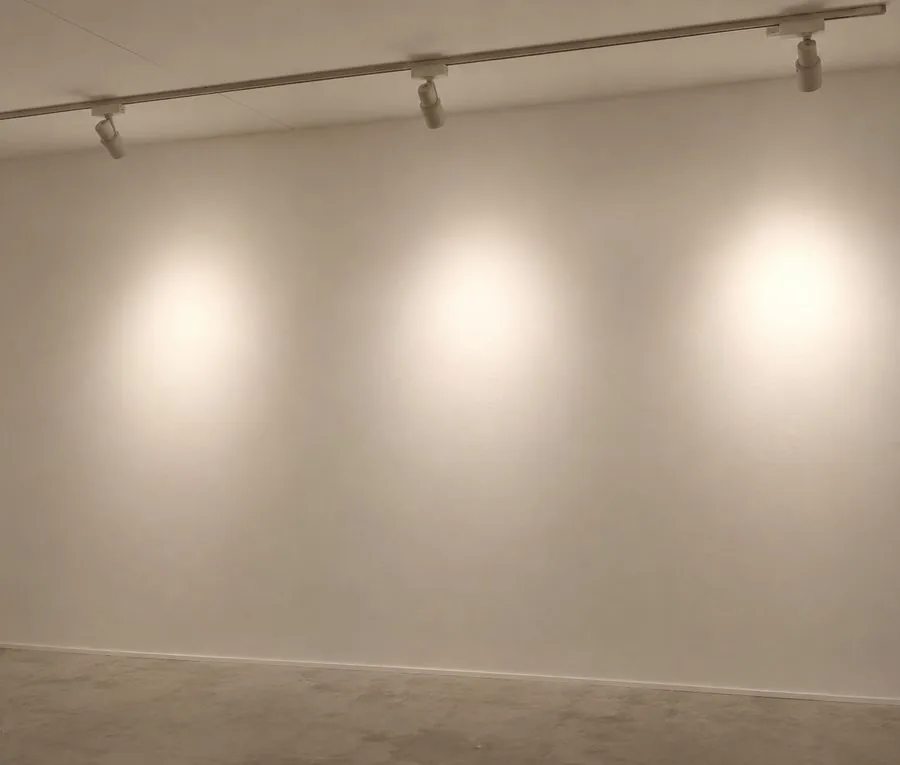 A large, completely blank wall in an empty room, washed by three track spotlights that pool light across the bare surface