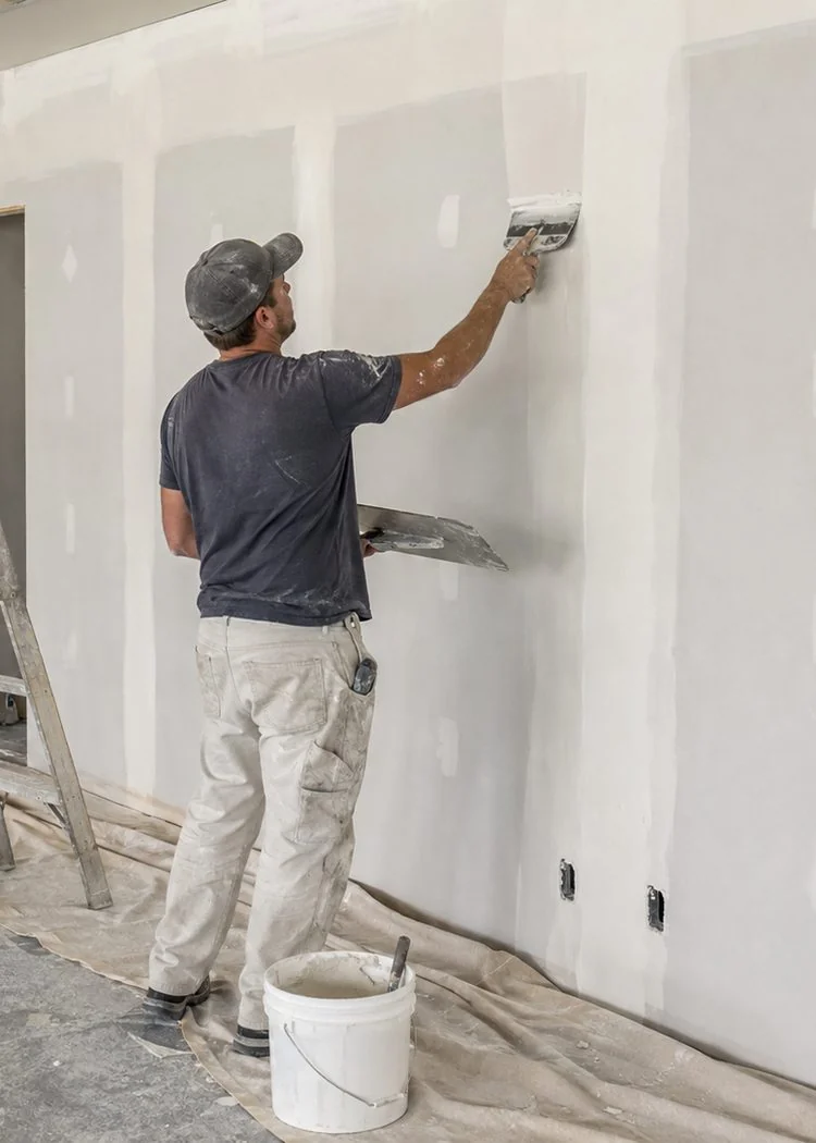 Tradesman skim-coating and finishing a drywall partition with a taping knife, floors covered and a ladder alongside, preparing the surface for printing